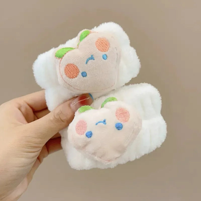Mãos segurando dois scrunchies peludos brancos com detalhes bordados em formato de rostos coloridos