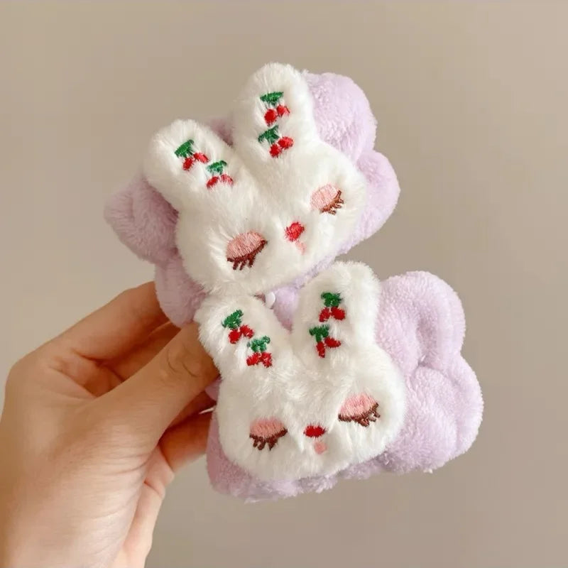 Mão segurando par de presilhas de cabelo lilás com coelhos de pelúcia branca bordados com cerejas vermelhas.