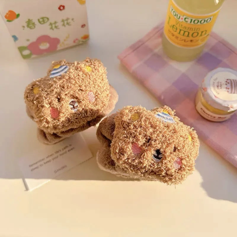 Meias felpudas marrom com rosto de ursinho e chapéu, sobre mesa com bebidas e papel decorativo