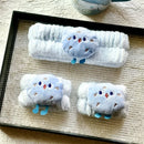 Faixa e polainas felpudas brancas com detalhe azul decorativo, exibidas em bandeja preta sobre superfície texturizada